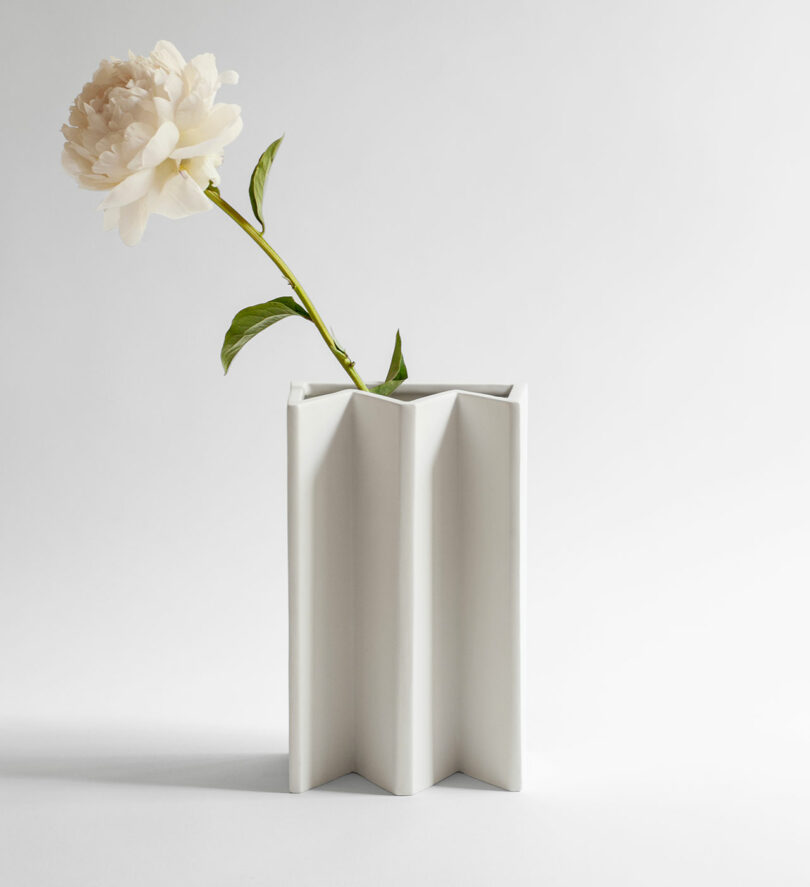 The white, angular ceramic vase holds a single peony flower and green stem, reminiscent of the minimalist elegance seen in Kish O Mat Sara Khamisi designs, set against a plain light background.