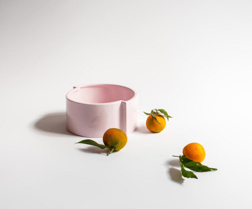 Pink ceramic bowl on a white surface with three small oranges, each with green leaves, scattered nearby.