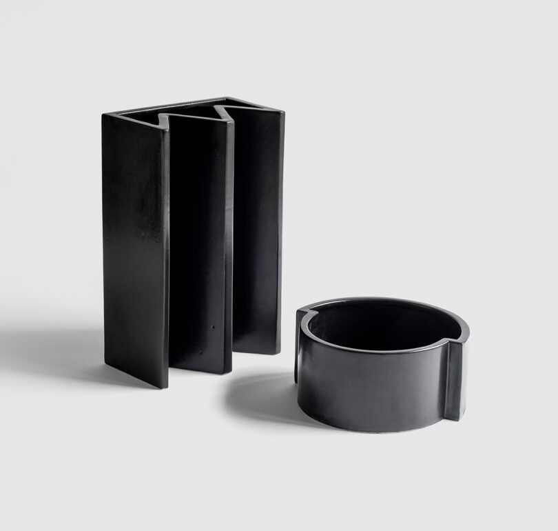 Two black, abstract-shaped sculptures on a plain gray background. One is a tall, rectangular block with slits; the other is a shorter, rounded structure with an opening.