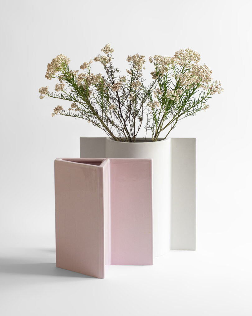 White vase with delicate white flowers and greenery, accompanied by an abstract pink ceramic sculpture on a white background.