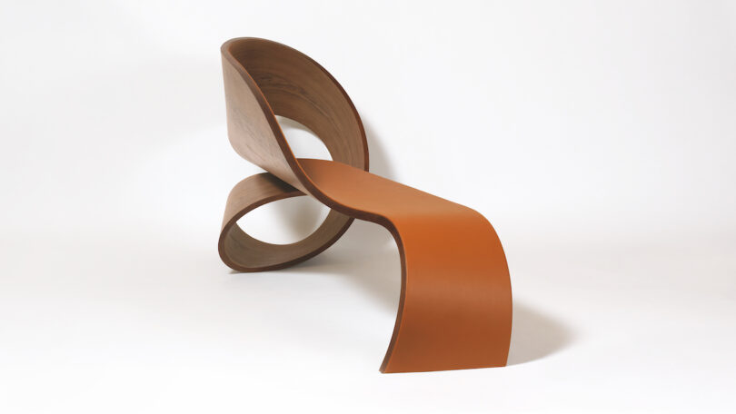 A modern, sculptural chair with a wooden looped backrest and a smooth, curving brown seat against a white background