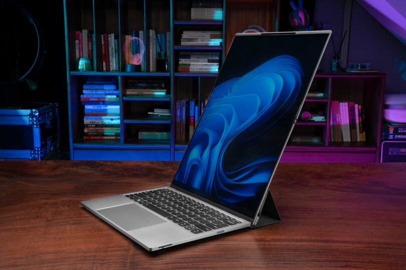 A sleek ThinkBook Flip rests on a wooden desk, displaying a blue abstract pattern on its screen, with shelves of books and tech items softly blurred in the background.