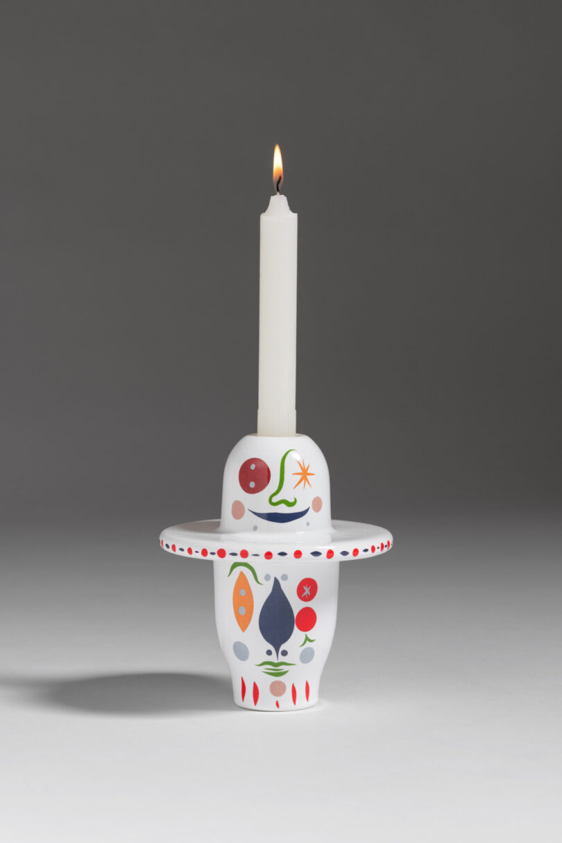 A lit white candle is inserted into a colorful, decorated ceramic holder with abstract patterns, set against a gray background.
