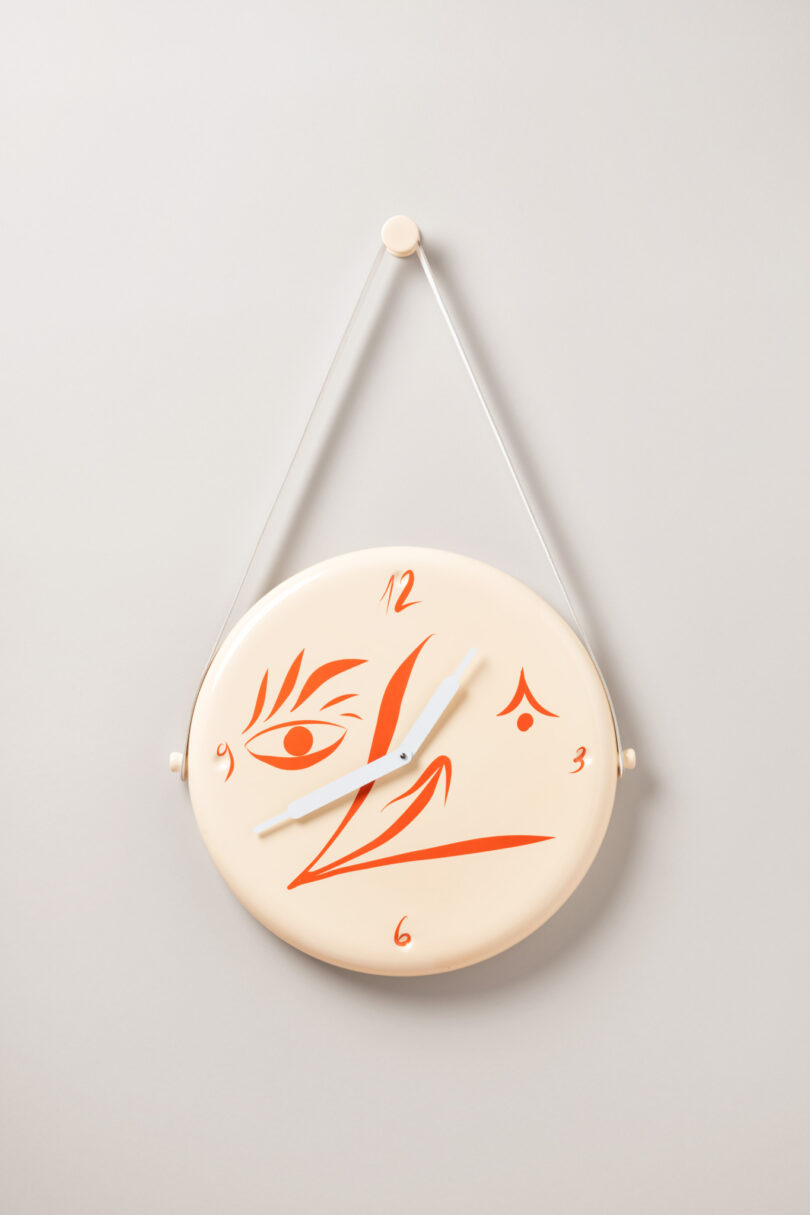 A round wall clock with red, abstract face features as decoration. The hour and minute hands are white, pointing to approximately 10:10. The clock hangs by a white string.