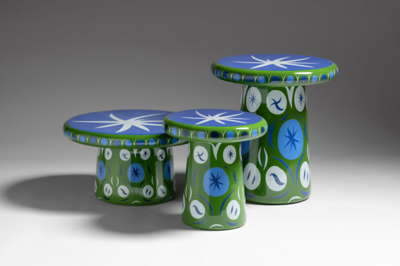Three colorful, mushroom-shaped stools with blue tops and green bases featuring white and blue abstract designs, set against a plain gray background.