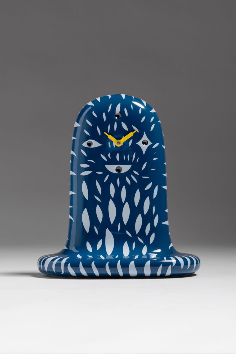 Blue and white stylized clock with abstract eyes and leaf patterns, featuring yellow hands, set against a plain gray background.