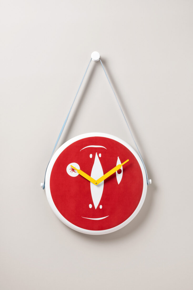 A red oval wall clock with abstract white and yellow designs, featuring yellow hour and minute hands, hangs by a thin strap on a beige wall.