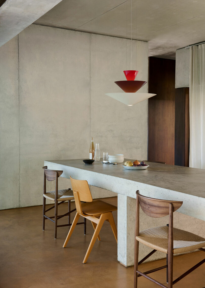 Modern dining space with a concrete table, three wooden chairs, and a geometric pendant light. The table has a glass carafe, plates, and fruit. Minimalist decor with neutral tones.