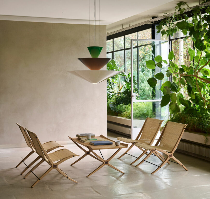 A minimalist room features four wooden lounge chairs and a low table with books, adjacent to large windows with lush green plants outside and inside.