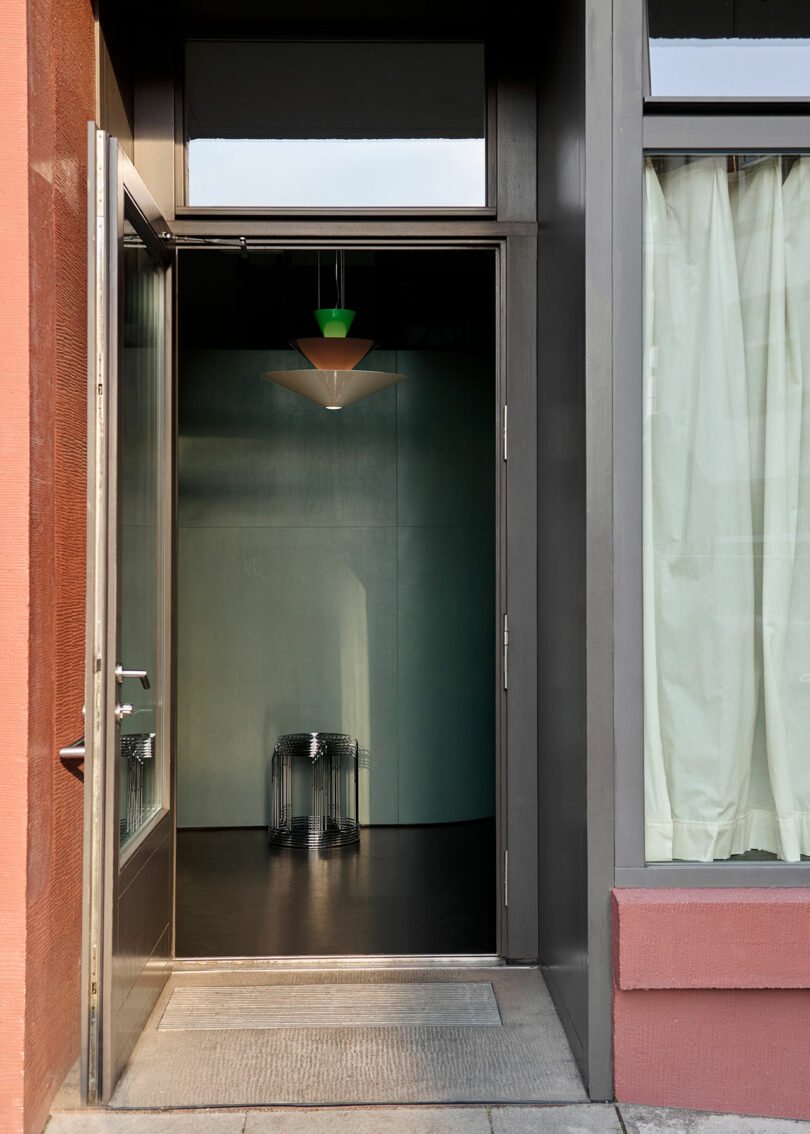 Open door to a dimly lit room with a small table, featuring a hanging light above. Curtain on the right and brick wall on the left.
