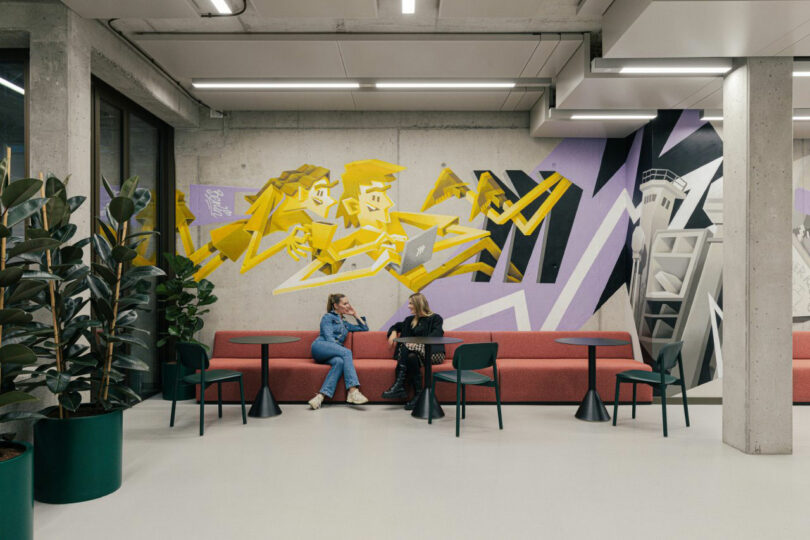 Two people sit on a red sofa in a modern space with graffiti art and plants. The room has tables, chairs, and overhead lighting.