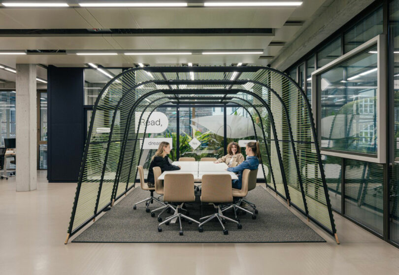 Four people sit around a table inside a transparent, tunnel-like meeting pod in a modern office space.