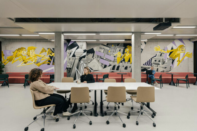 Three people are seated in a modern meeting room with mural art on the walls, featuring abstract yellow figures and geometric shapes.