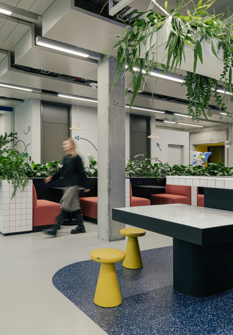 Modern office space with red seating, plant decor, and yellow stools. A person in motion blurs past a concrete pillar. Blue speckled flooring contrasts with minimalist furnishings.