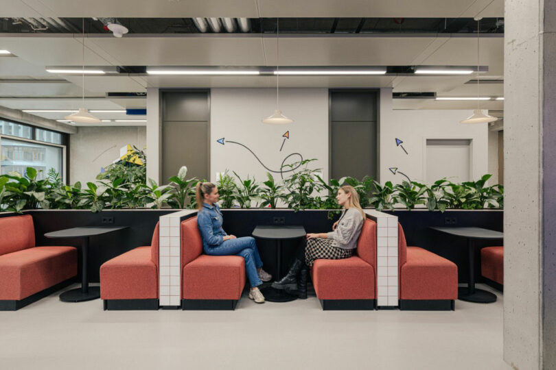 Two people sitting on red booths face each other in a modern office space, with green plants and wall art in the background.