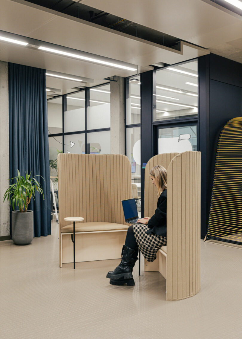 A person seated on a booth sofa using a laptop in a modern office space with plants and glass partitions.