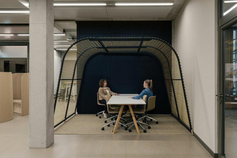 Two people sit at a rectangular table in a modern office space, enclosed by an arched structure with wooden slats.