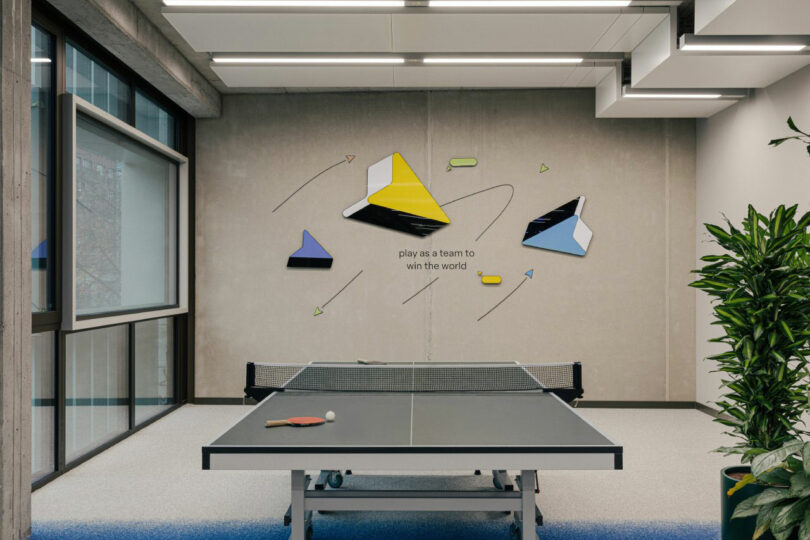 A ping pong table with paddles is in a modern room. Geometric wall art and the phrase 
