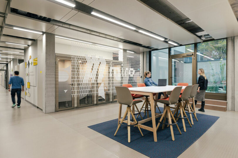 Modern indoor office space with a high table and chairs, abstract wall art, and large windows. Three people are present, two sitting and one standing.