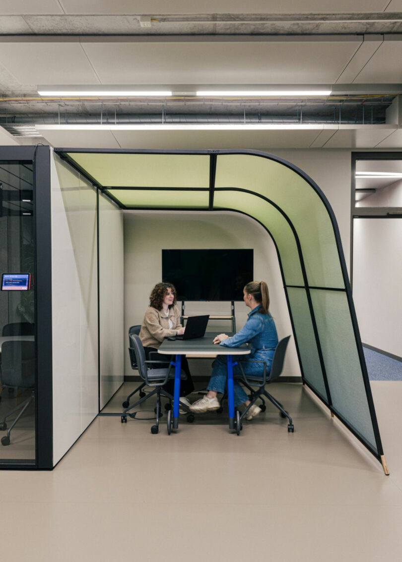 Two people are seated at a small table in a modern, semi-enclosed office space with glass walls. One person uses a laptop. A television is mounted on the back wall.