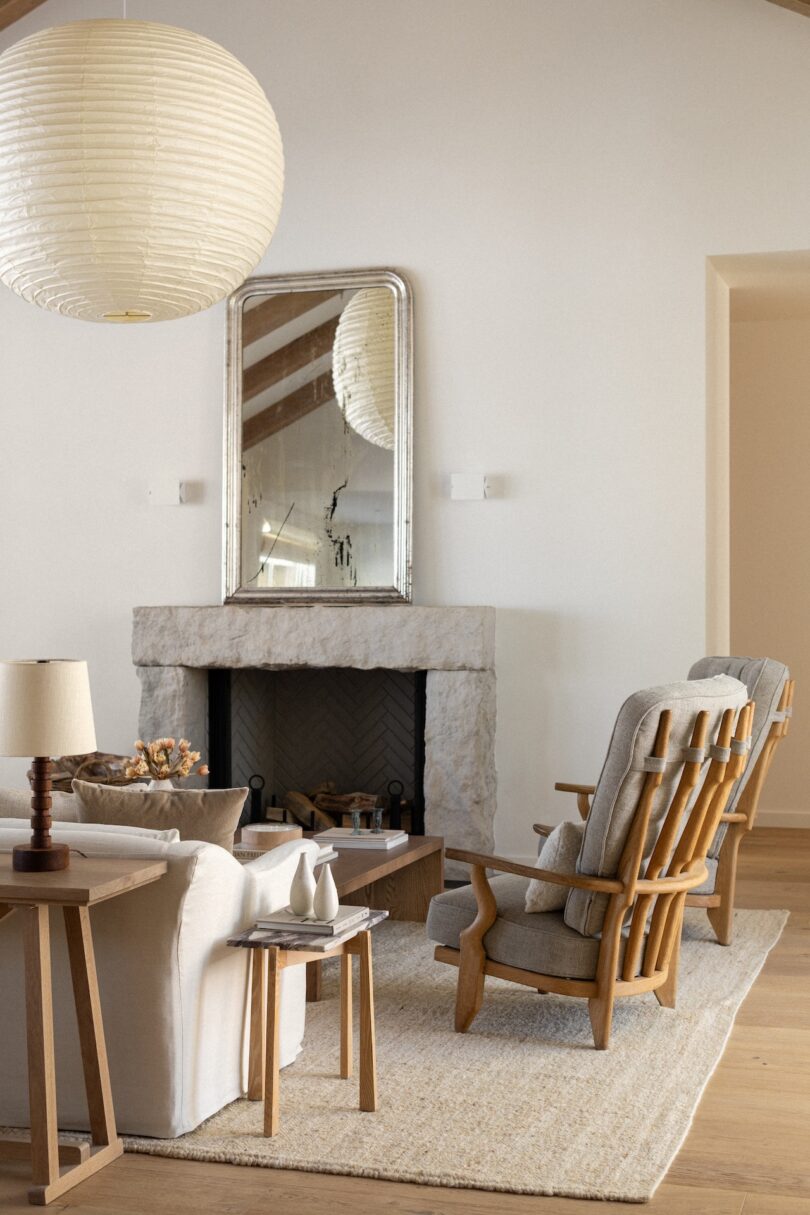A cozy living room with a fireplace, a large mirror above it, a white sofa, two wooden chairs, a side table with a lamp, and a large round ceiling light. Beige and wood tones dominate the decor.
