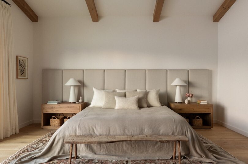 A modern bedroom with a large bed, beige bedding, wooden nightstands, two lamps, a bench at the foot, and a rug. Ceiling features wooden beams and walls are painted white.