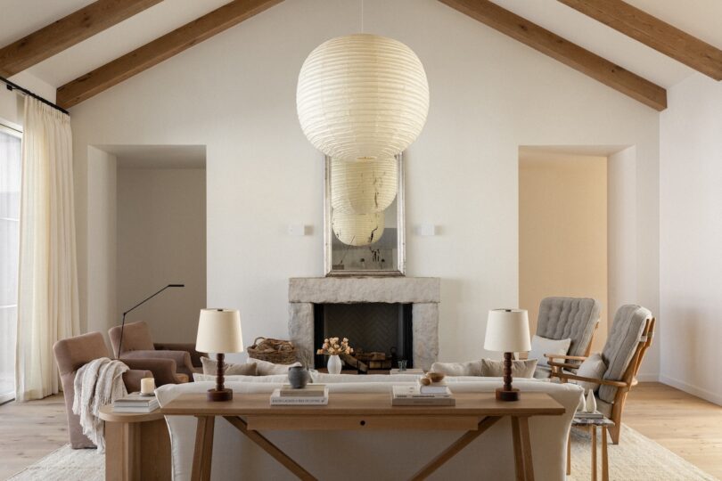 Spacious living room with high ceilings, wooden beams, a central fireplace, and modern furniture. A large round pendant light hangs above a table with lamps and decorative items.