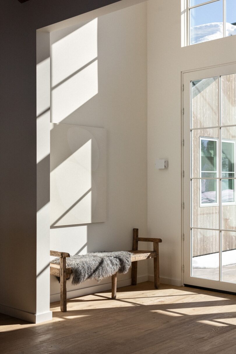 A sunlit corner with a wooden bench covered by a furry throw near a large window. Shadows cast on the wall and floor.
