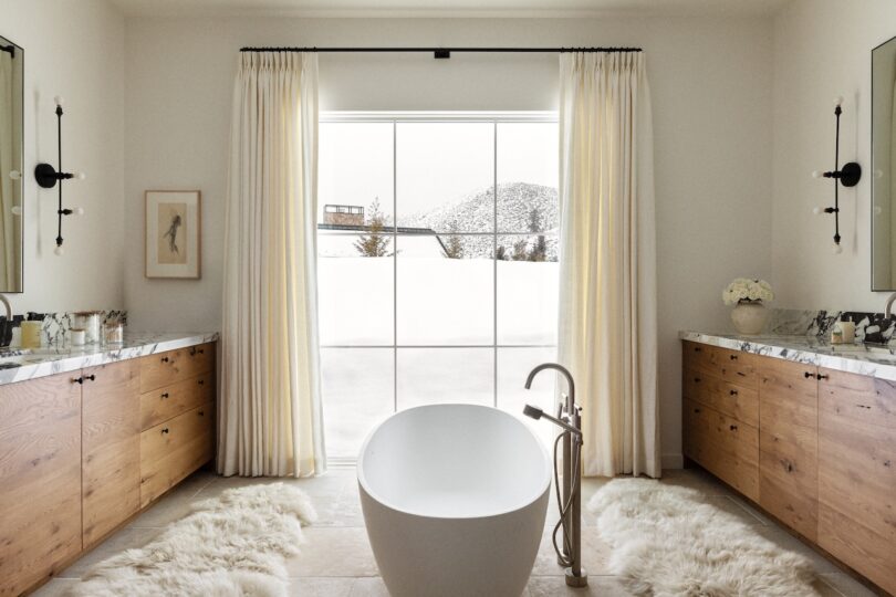 Modern bathroom with a freestanding tub, wooden vanities on both sides, and large window with sheer curtains. Two wall sconces flank a simple artwork, and plush rugs are on the floor.