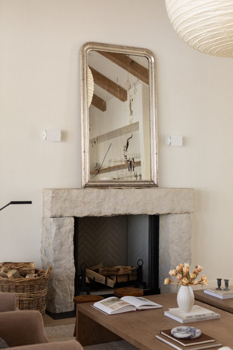 Living room with a stone fireplace, large mirror above, wicker basket, wooden coffee table with books, and a vase of flowers.