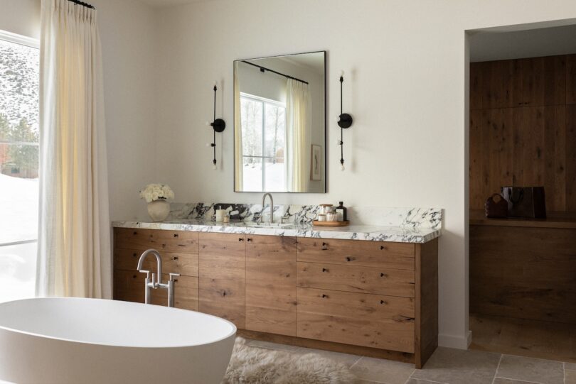 Bathroom with a wooden vanity, marble countertop, and large mirror. Freestanding bathtub and light curtains by a window are visible. Minimalist decor with natural tones.