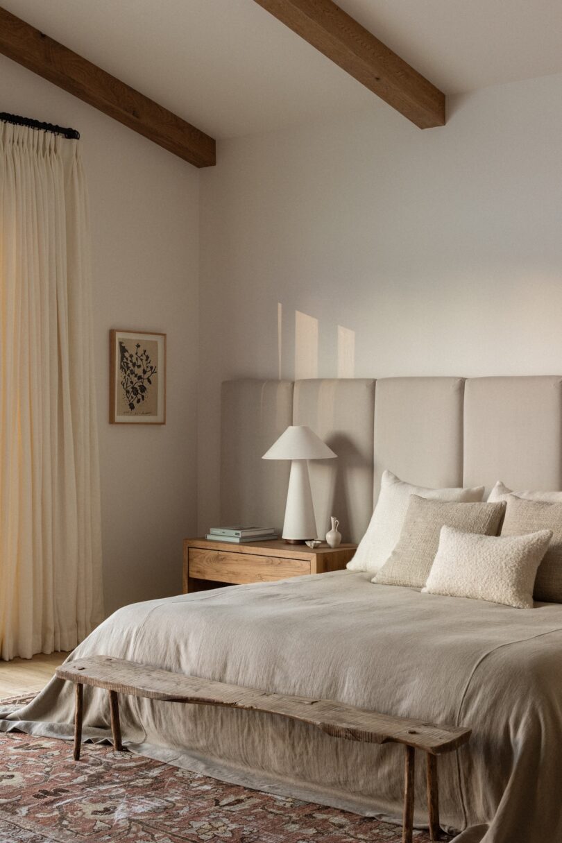 A cozy bedroom with a beige bed, wooden bench, and nightstand. A lamp and book sit on the nightstand. Light filters through a window with cream curtains. Exposed wooden beams on the ceiling.