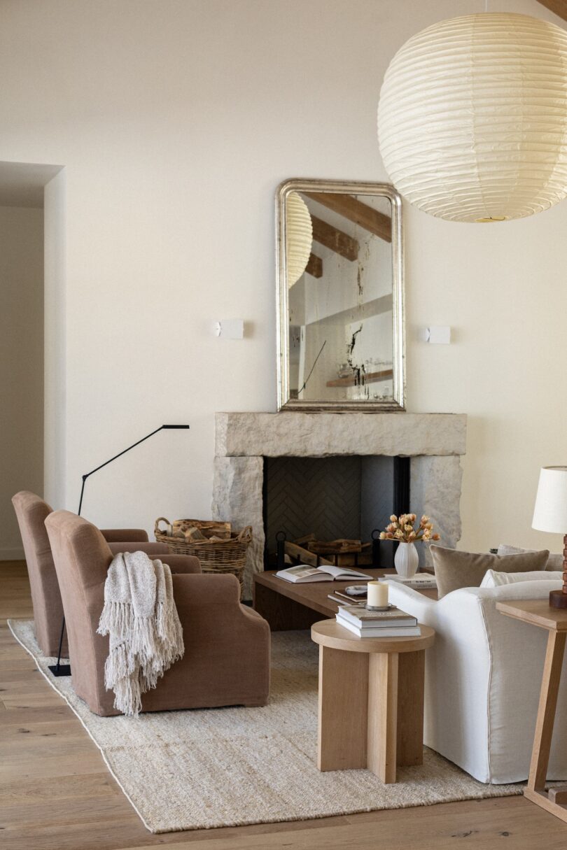 A cozy living room with a stone fireplace, a large mirror above it, two brown armchairs, a white armchair, a wooden coffee table, and a round paper lantern hanging from the ceiling.