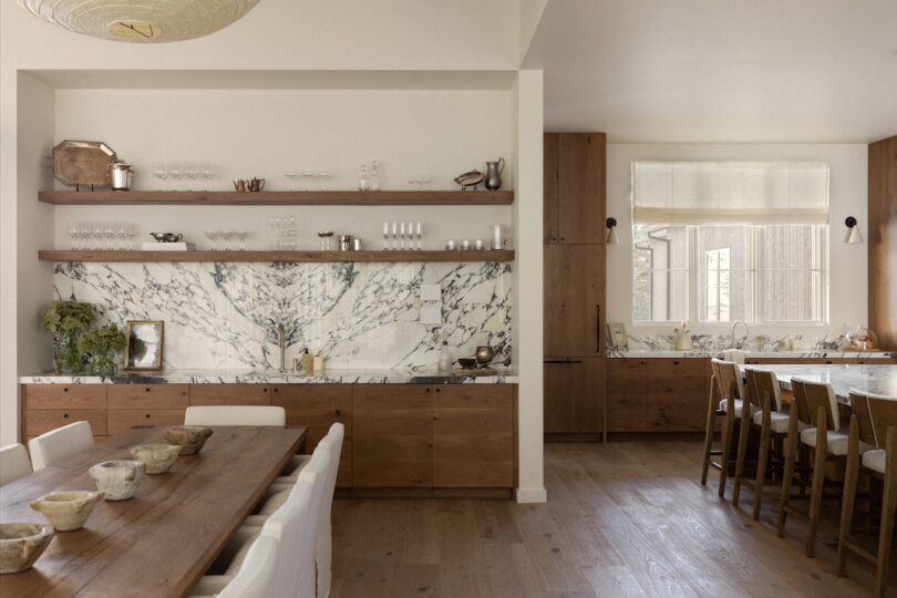 Modern kitchen with wooden cabinets and marble countertops. Open shelving displays glassware. Long dining table with chairs on the left, large window and bar chairs on the right.