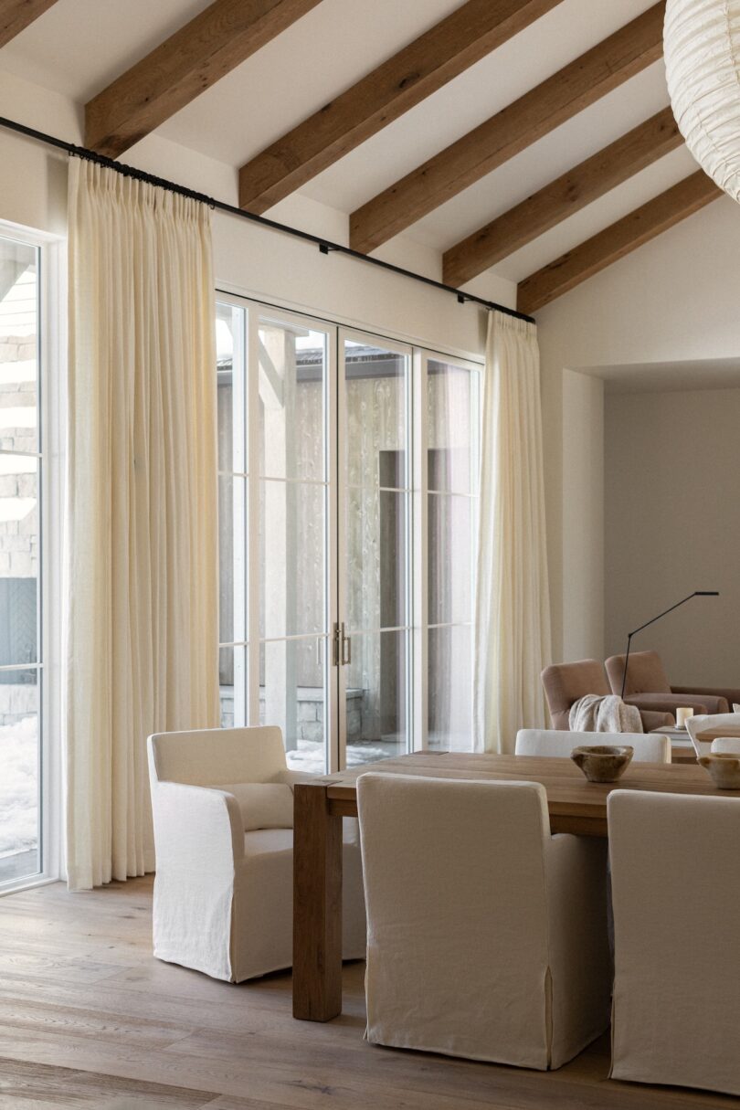 Room with high ceiling, wooden beams, large windows with curtains, and a table surrounded by white upholstered chairs. Natural light fills the space.