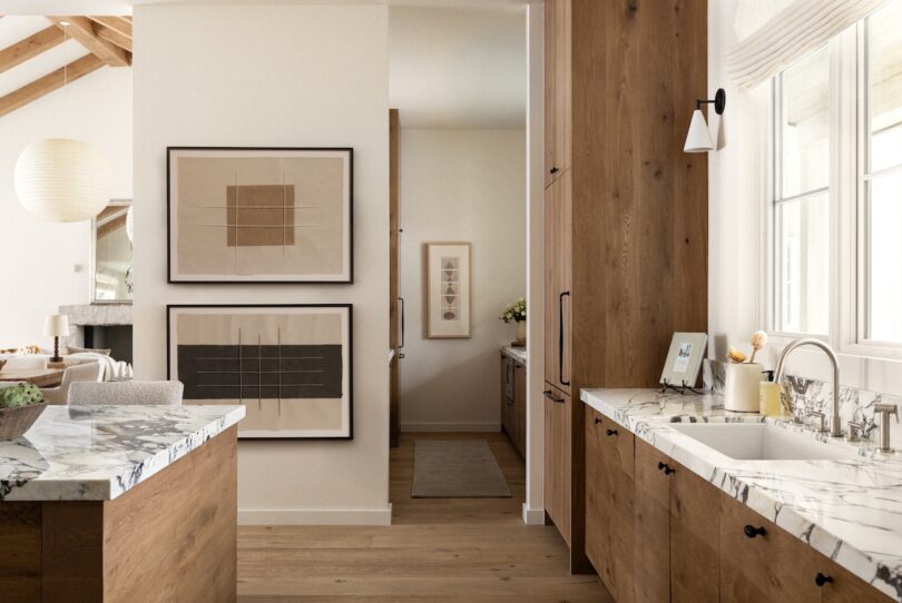 Modern kitchen with wooden cabinets, marble countertops, and a sink by a window. Framed artworks hang on a wall, and a glimpse of an open living area with a hanging lamp is visible.