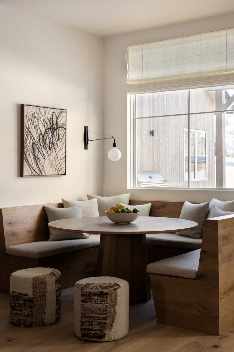 Cozy corner with a round wooden table, cushioned bench seating, abstract wall art, and a bowl of fruit. Large window with a view and two textured stools complete the setup.