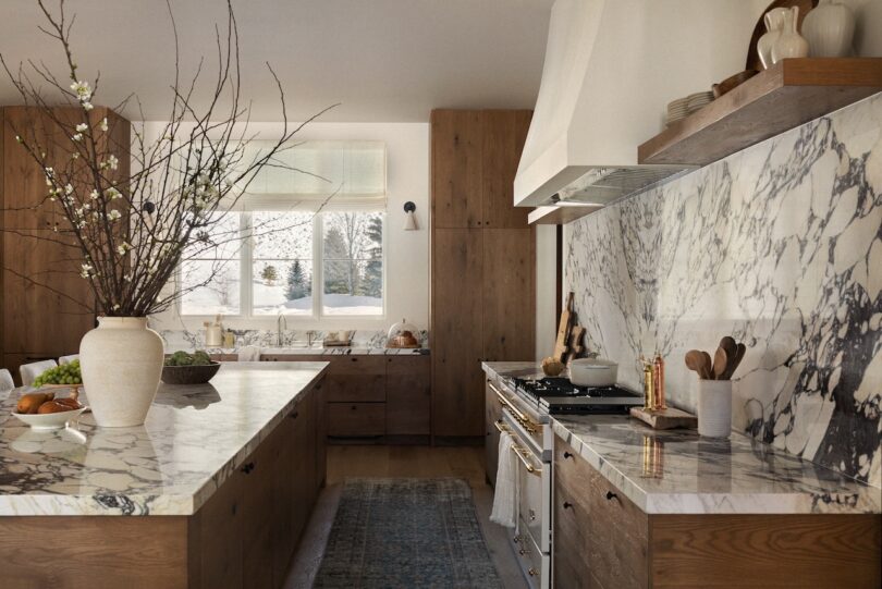 Modern kitchen with marble countertops, wooden cabinets, and an island. A vase with branches stands on the island. Large window offers a snowy outdoor view.