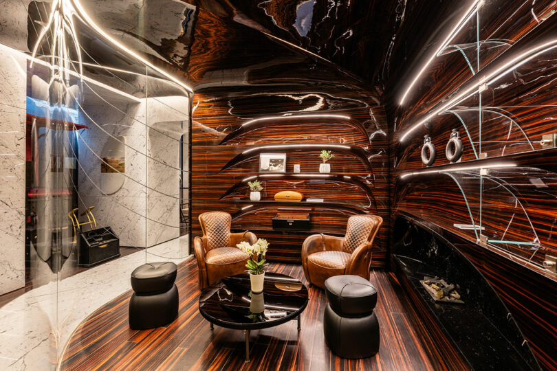 Luxurious room with two leather chairs, a black coffee table, and curved wood-paneled walls with shelves holding decorative items. Marble accents and modern lighting enhance the design.