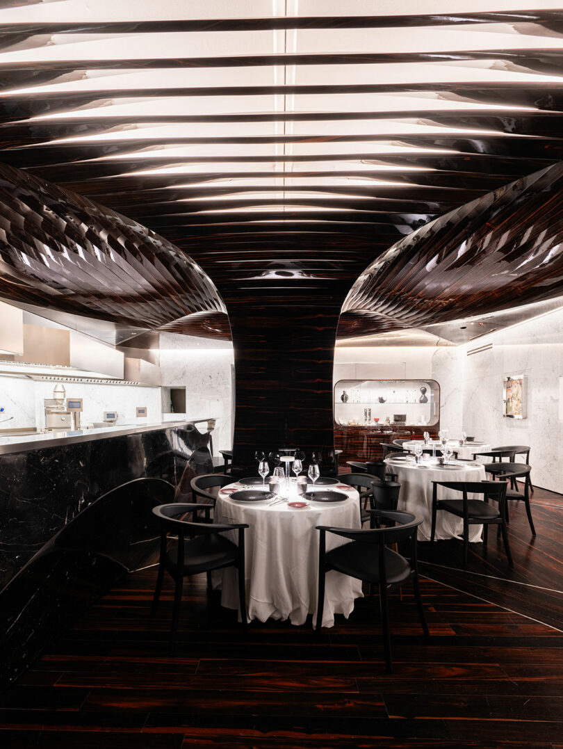 Modern restaurant interior with dark, wooden ceiling and floor. Round tables with white tablecloths and black chairs are set elegantly. An open kitchen is in the background.