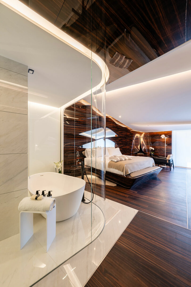 Modern bedroom with a curved glass partition separating a freestanding bathtub and wooden interiors, featuring a bed, wooden floor, and soft lighting.
