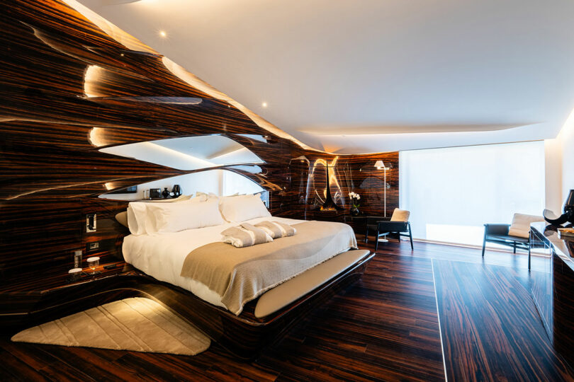Spacious modern bedroom with sleek wooden walls and flooring, a large bed with white bedding, contemporary furniture, and ambient lighting. Large window with a sheer curtain.
