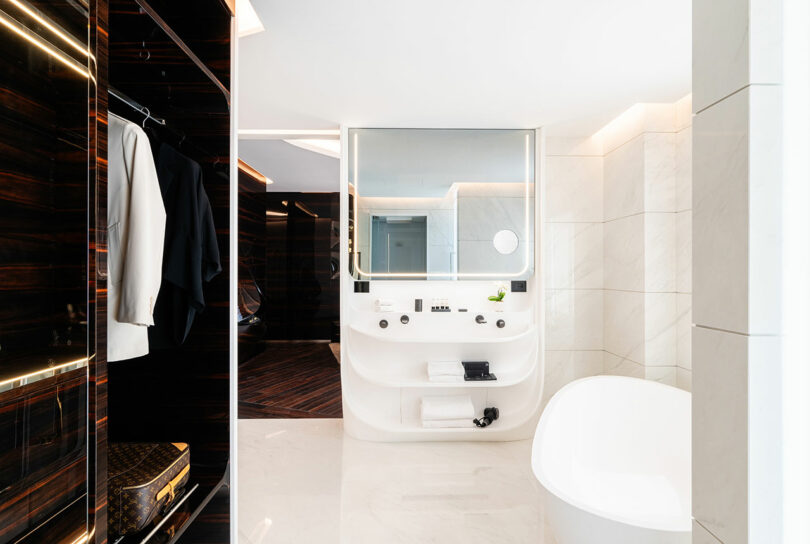 Modern bathroom with a large white bathtub, wall mirror, sink, and a closet with hanging clothes. The design features sleek surfaces and a minimalist aesthetic.