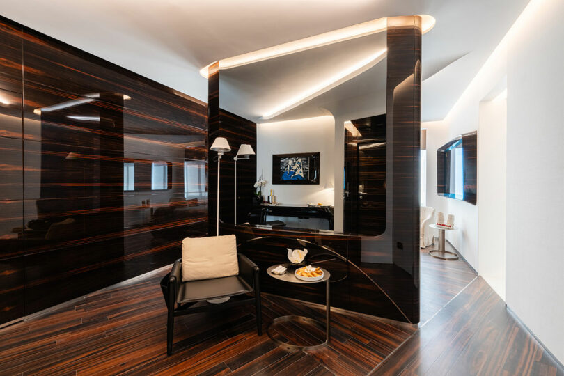 Modern interior featuring dark wood paneling, a black chair with a beige cushion, a small round table with plates, and a wall-mounted TV in a sleek, reflective environment.