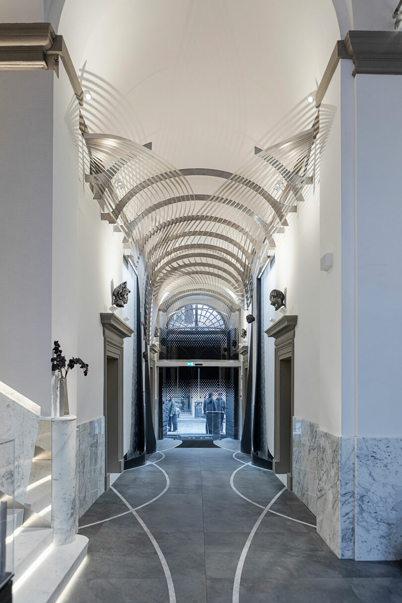 Modern hallway with marble walls and geometric ceiling design, leading to an arched glass entrance. Floor features curved line patterns.