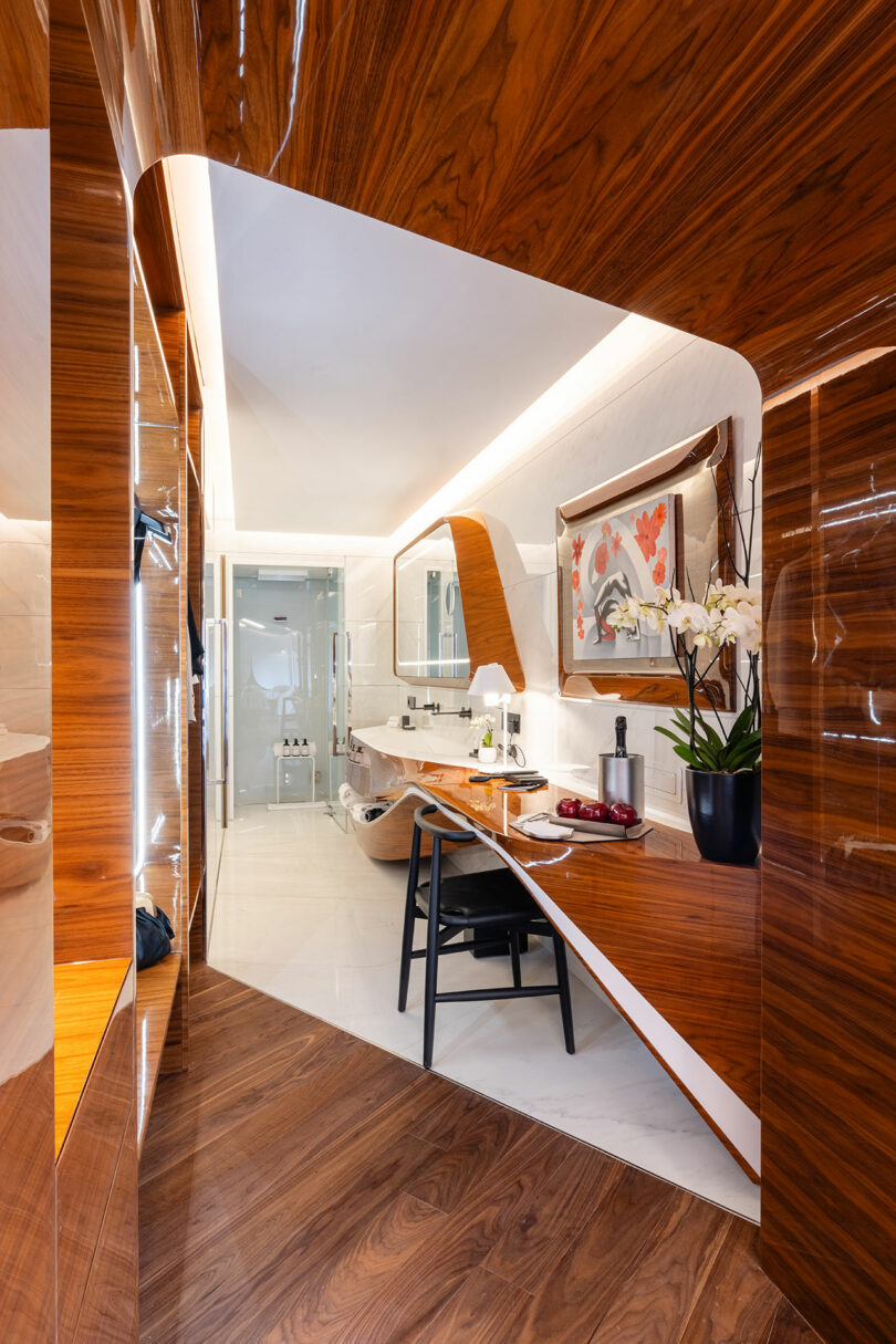 Modern bathroom with wooden paneling, a white freestanding tub, glass-enclosed shower, and a vanity with a chair. A potted orchid and artwork are on the counter.