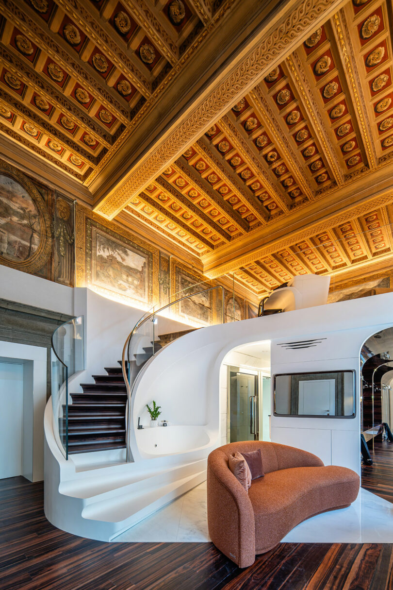 Modern interior with ornate coffered ceiling, curved staircase, and a brown sofa. The room blends classical and contemporary design elements.