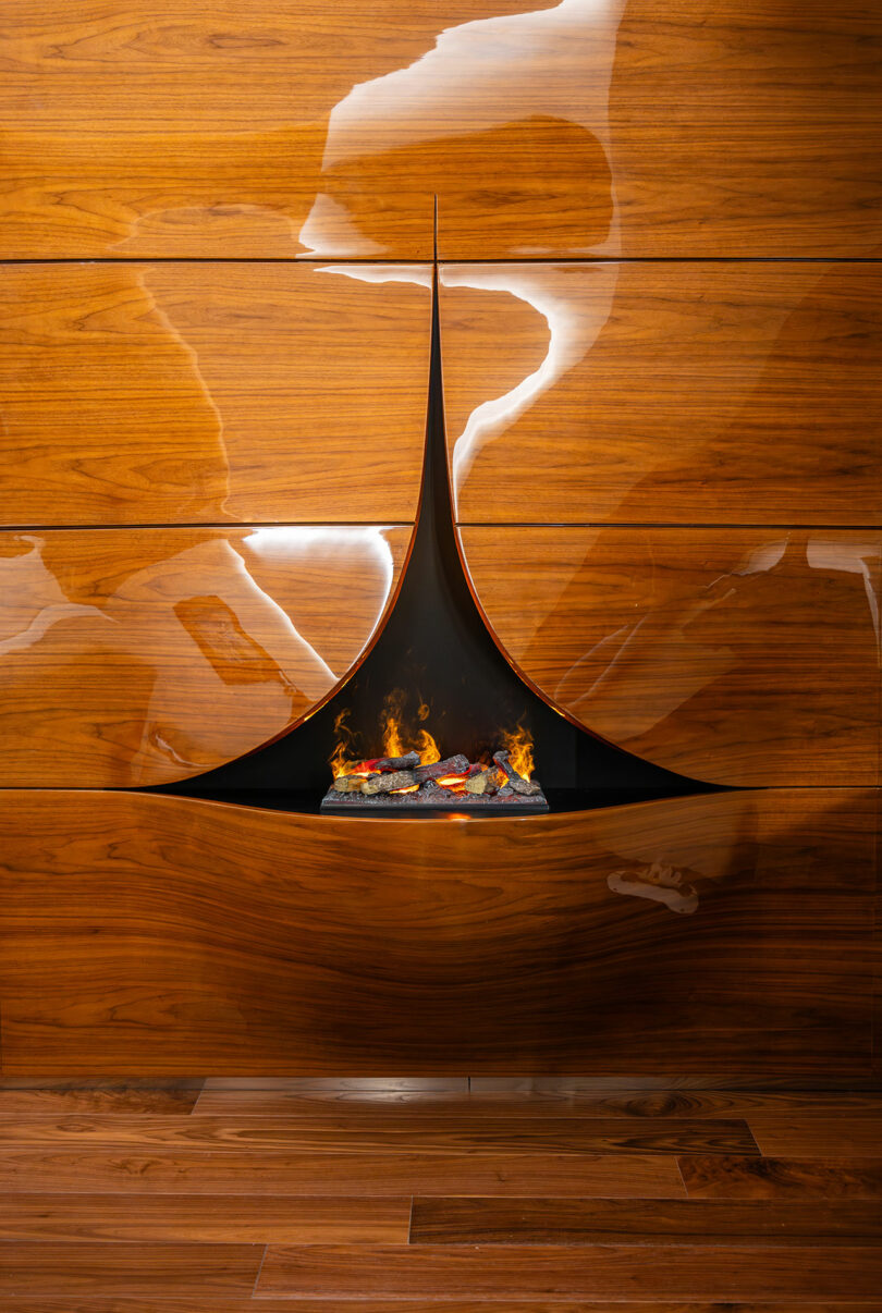 Modern wall-mounted fireplace with a sleek, curved black hood against glossy wood paneling, emitting a small flame with logs.