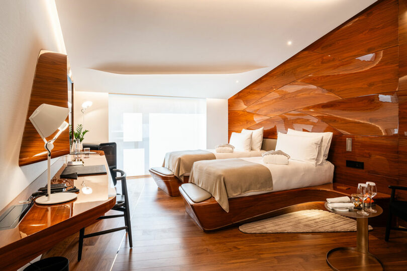 Modern hotel room with two twin beds, glossy wood paneling, and a sleek desk. A small table holds champagne glasses, and natural light filters through a large window with sheer curtains.