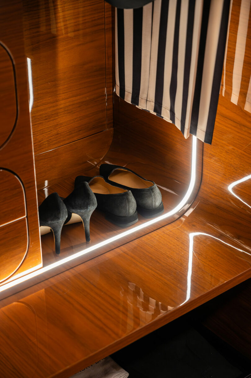 A pair of black high heels on a glossy wooden shelf with underlighting. Above the shoes, a black and white striped fabric hangs.